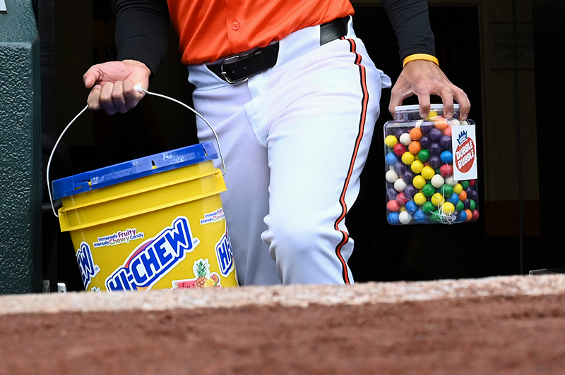 A tight shot of an Orioles bat boy climbing up the dugout steps. You can only see from his belt to just below his knees, but he's carrying a plastic container of colorful Dubble Bubble gumballs in his left hand and an enormous yellow Hi-Chew bucket in his right hand. It looks heavy. It's the huge size that your coach would store baseballs in.