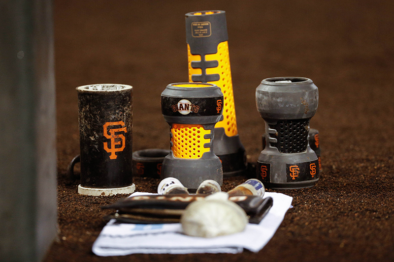 A picture of four different kinds of batting weight sleeves, all with Giants logos on them, arranged standing up on the dirt.