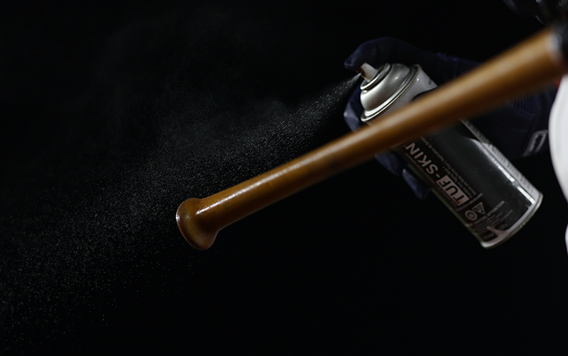 A closeup of a hand in a batting glove spraying a fine mist of Cramer Tuf-Skin onto the handle of a bat.